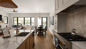 A modern kitchen with a large island, a dining area with a table and chairs, and a living room with a sofa and large windows. - Luxury 3-Story Lakehouse |Pool + Hot Tub + Sundeck (Horseshoe Bay)