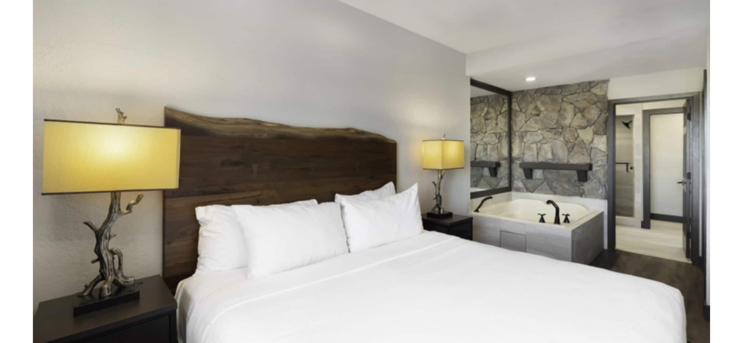 Een hotelkamer met een groot bed, twee bedlampen, een stenen muur, een badkuip en een deur naar een andere kamer.
