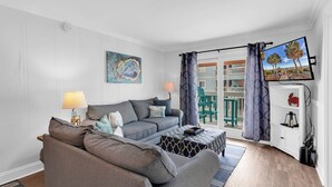 Ein Wohnzimmer mit Sofa, Couchtisch, einem Gemälde, einem Fernseher und einem Balkon mit Gartenmöbeln.