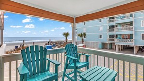 Ein Balkon mit zwei Stühlen und einem Tisch, der Blick auf einen Strand und ein Gebäude mit mehreren Balkonen bietet.
