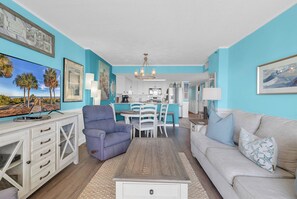 A living room with a TV, a sofa, a dining area with chairs, and a kitchen in the background. - Atalaya Towers 1004 - Oceanfront in Garden City Beach (Garden City Beach)