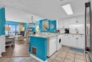 A modern kitchen with white cabinets, a central island, a built-in washer and dryer, and a large window overlooking a balcony. - Atalaya Towers 1004 - Oceanfront in Garden City Beach (Garden City Beach)