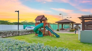 Un parque infantil con un tobogán y una estructura para escalar, rodeado por una zona cercada que incluye una mesa de picnic y un pabellón.