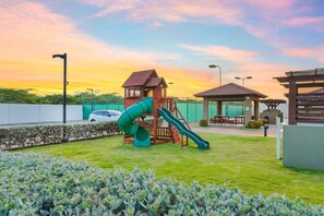 A playground with a slide and climbing structure, surrounded by a fenced area with a picnic table and a gazebo. - Beautiful 2BR2BA with Great Amenities @ PalmBeach (Noord)
