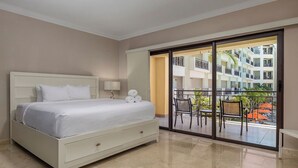Habitación de hotel con cama, mesita de noche, lámpara y puerta corrediza de vidrio a un balcón con mobiliario exterior.