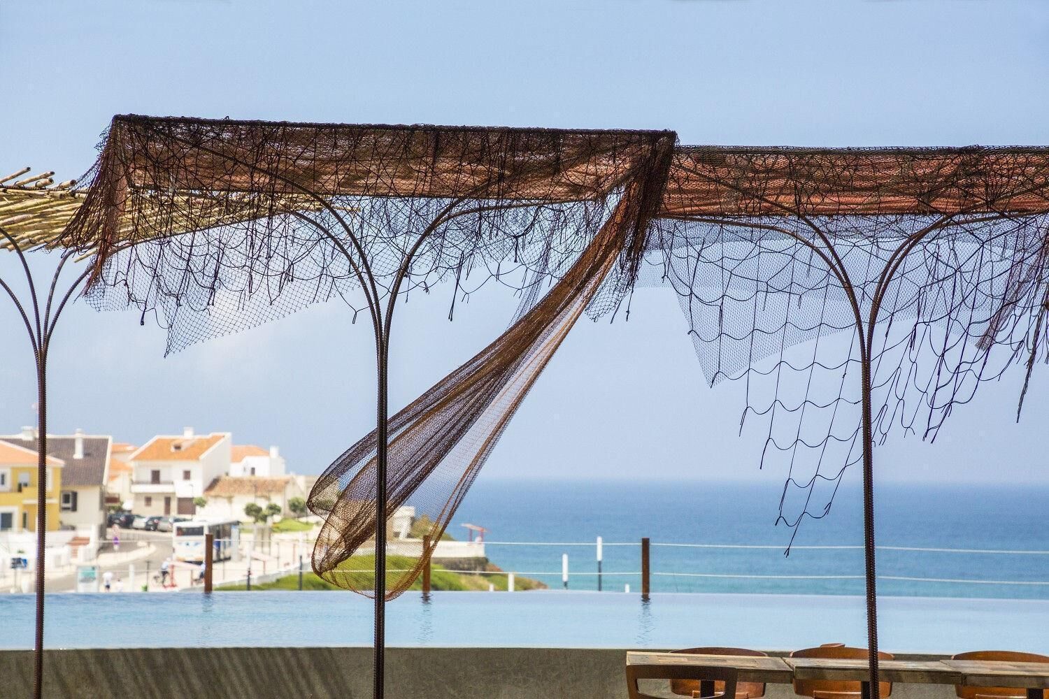 A seaside view with a pool, a netted pergola, and buildings in the background.