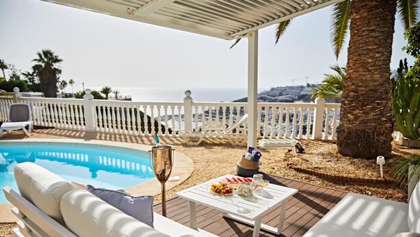 Ein Poolbereich mit weißer Sitzgelegenheit im Freien, einem Tisch mit Speisen und Getränken und Blick aufs Meer.