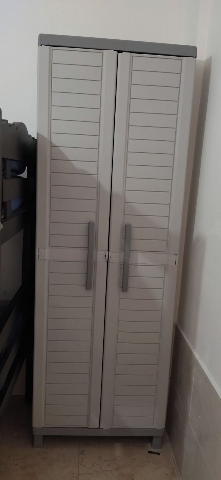 A grey wardrobe with two doors and handles, located in a corner of a room with tiled flooring.