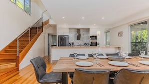 A modern kitchen with a central island, a dining area with a wooden table and leather chairs, and a staircase leading to an upper level. - Macadamia Ocean Escape (Pottsville)