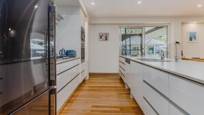 A modern kitchen with a built-in refrigerator, wooden flooring, white cabinetry, and a view of the outside through a large window. - Macadamia Ocean Escape (Pottsville)