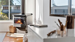 Eine moderne Küche mit Kaminofen, weißer Arbeitsplatte und Blick ins Freie durch ein großes Fenster.
