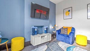 A modern living room with a blue sofa, yellow ottoman, white entertainment unit, and a mounted TV displaying 'NETFLIX'. - Gorgeous 2BR flat in Fulham & Chelsea (London)