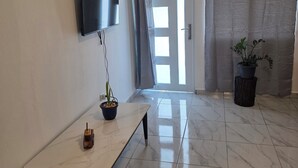 Una habitación con una televisión de pantalla plana montada en la pared, una mesa pequeña con una planta en maceta, una puerta con cortinas y otra planta en maceta sobre un taburete de madera.