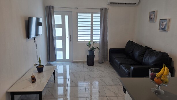 Una sala de estar con televisor, un sofá de cuero negro, una mesa de comedor y una planta en maceta.