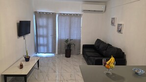 Una sala de estar con un sofá, un televisor montado en la pared, una planta en maceta y una mesa con fruta.