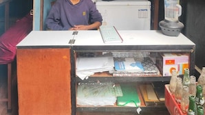 A small, cluttered room with a person sitting at a desk, surrounded by various items including a water dispenser, a coffee machine, and a refrigerator. - Anand Home Stay (Kathmandu)