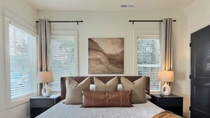 Une chambre à coucher avec un grand lit, deux tables de chevet, une tête de lit, une œuvre d’art accrochée au mur et une fenêtre avec des stores.