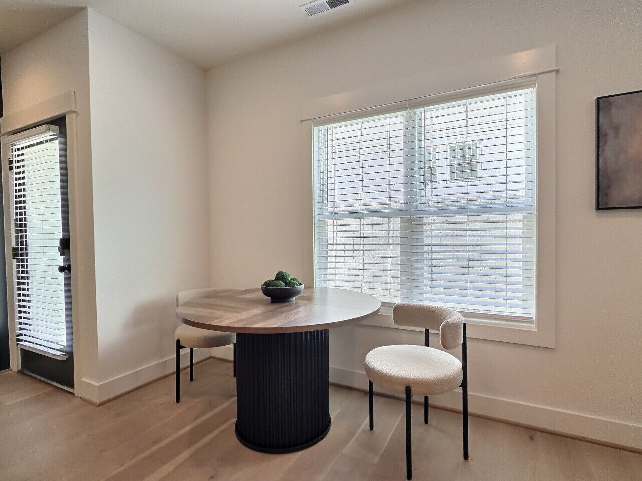 A dining area with a round table, two chairs, a bowl of fruit, and a window with blinds.