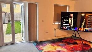 Ein Zimmer mit einem großen Flachbildfernseher auf einem Schrank, einer Glasschiebetür, die in einen Garten führt, und einem farbenfrohen Teppich.
