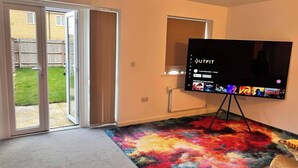 Ein Zimmer mit einem großen Flachbildfernseher auf einem Standfuß, einem bunten Teppich und einer Schiebetür zu einem Garten.