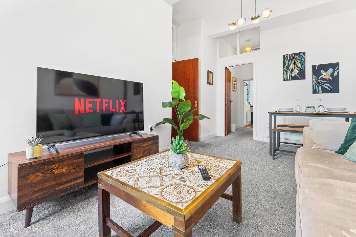 A living room with a flat-screen TV displaying 'NETFLIX,' a wooden coffee table with a decorative vase, a sofa, and a dining area with a table and chairs.