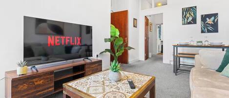 A living room with a flat-screen TV displaying 'NETFLIX,' a wooden coffee table with a decorative vase, a sofa, and a dining area with a table and chairs.
