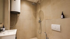 Un baño con lavamanos blanco, dispensador de jabón manual, cabezal de ducha de mano y calentador de pared.