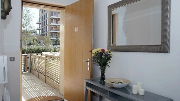 Vista interior de una habitación de hotel con puerta de madera, un espejo, un jarrón con flores y una mesita con un bol y velas.