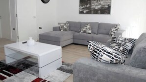 Una sala de estar moderna con un sofá seccional gris, una mesa de centro blanca, cojines estampados, una pintura de un paisaje urbano y un reloj de pared.
