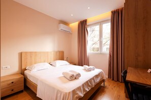 A hotel room with a wooden bed, a nightstand, a table, and a window with curtains. - Grandstay Tirana Central 01 (Tirana)