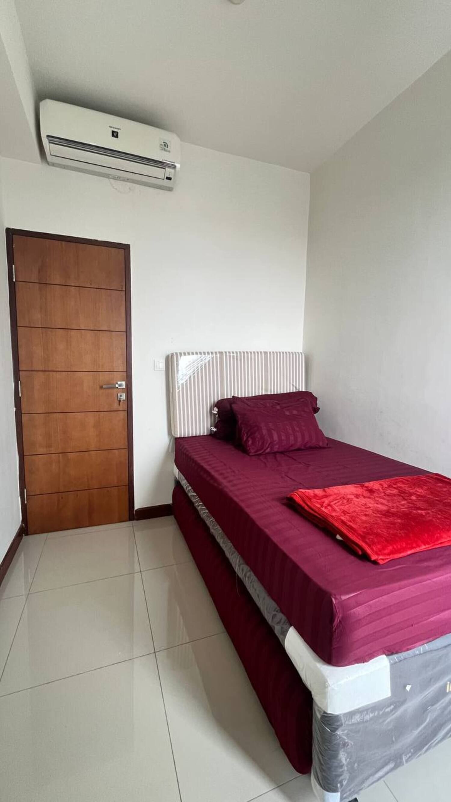 A small, single-bed room with a wooden door, a white air conditioner, and a red bedspread.