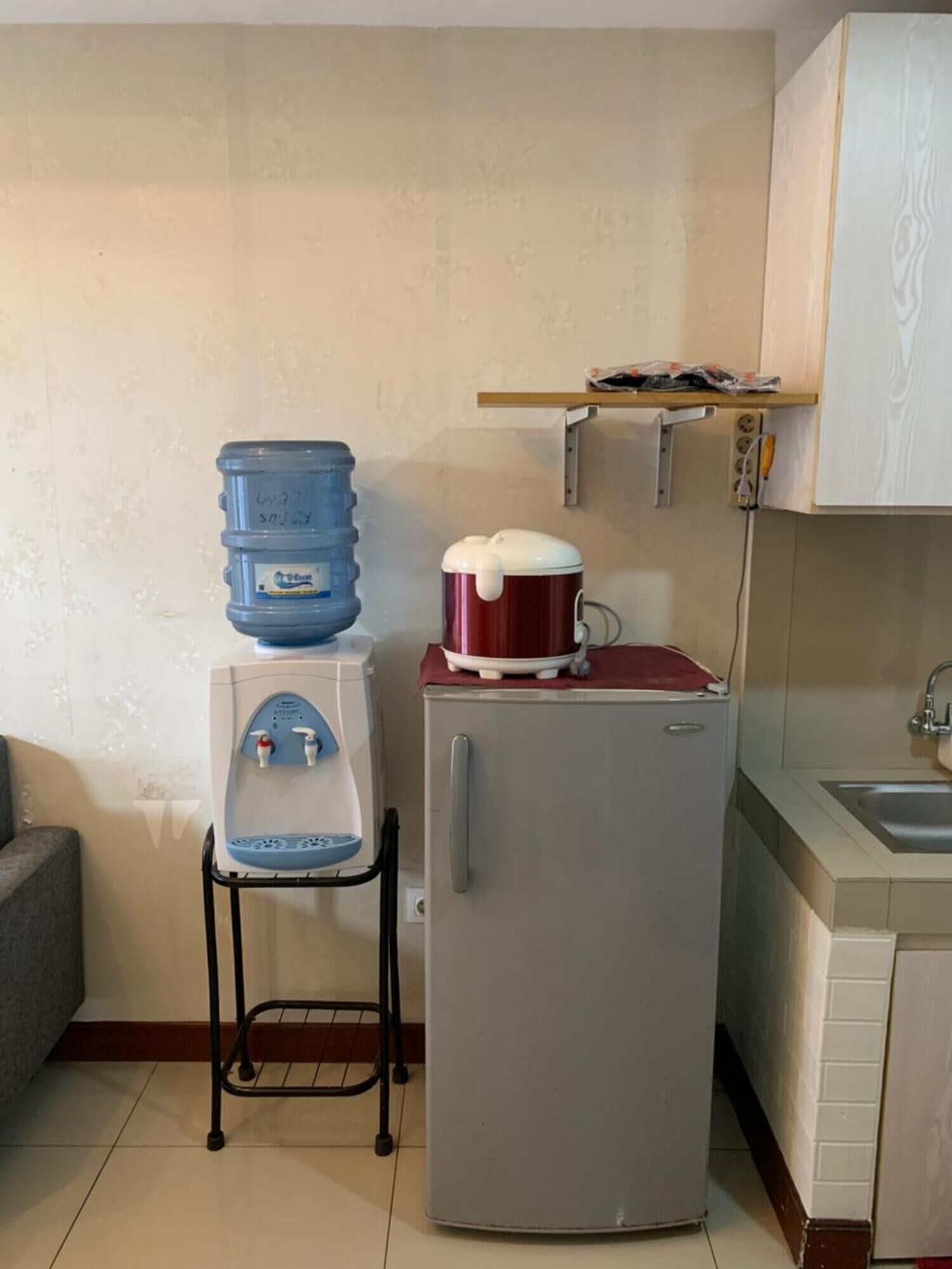 Een compacte keuken met een waterdispenser, een rode pan op een standaard, een koelkast en een spoelbak.