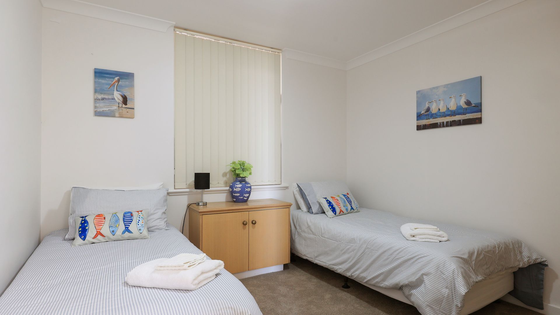 Une chambre avec deux lits simples, une commode en bois, une plante en pot et deux tableaux accrochés au mur.
