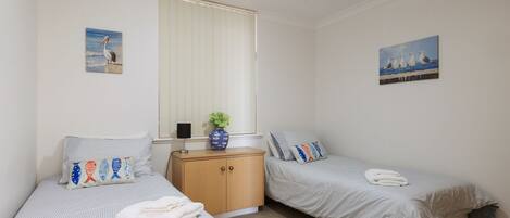 Une chambre avec deux lits simples, une commode en bois, une plante en pot et deux tableaux accrochés au mur.