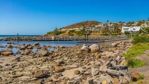 Una playa rocosa con grandes rocas, cielo azul despejado y una colina con edificios al fondo.