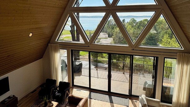 A spacious living area with a high ceiling, wooden beams, and large windows offering a view of a deck and lake. - Welcome to Torch Hill A-Frame, perched above iconic Torch Lake. (Alden)