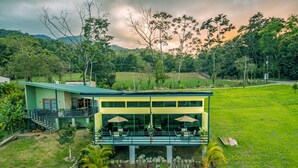 A modern two-story house with a balcony, surrounded by a lush garden and a hilly landscape. - The Honeybee House: Spacious 2BR Jungle Home (La Fortuna)