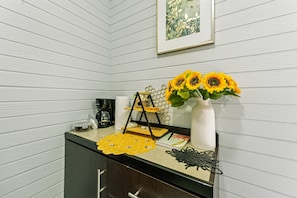 Un comptoir de cuisine avec une machine à café, un set de table à motifs en forme de nid d’abeilles, un vase avec des tournesols et un tableau encadré accroché au mur.