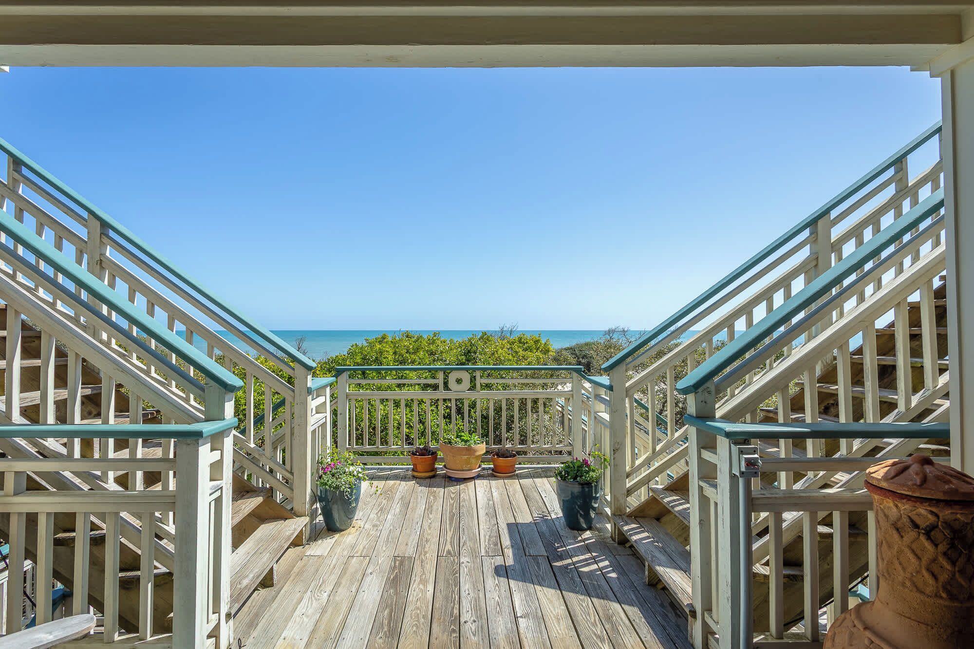 A wooden deck with a view of the ocean, potted plants, and a white railing.