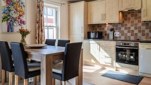 A modern kitchen with wooden cabinets, a dining table with chairs, a coffee machine, an oven, and a microwave. - Yr Encil - 2 Bedroom Apartment - Tenby (Tenby)