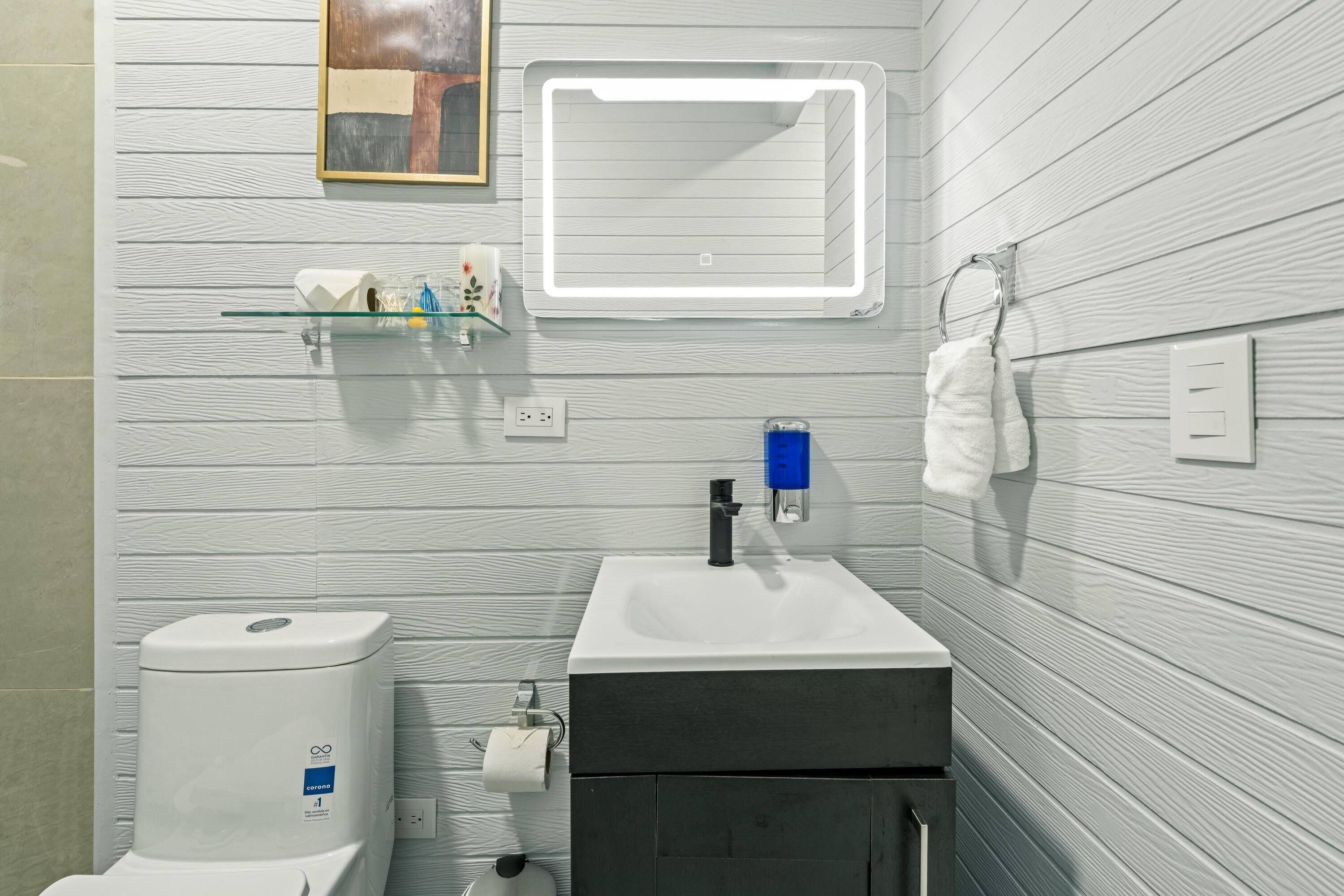 A bathroom with a toilet, sink, mirror, towel rack, and a shelf with toiletries.