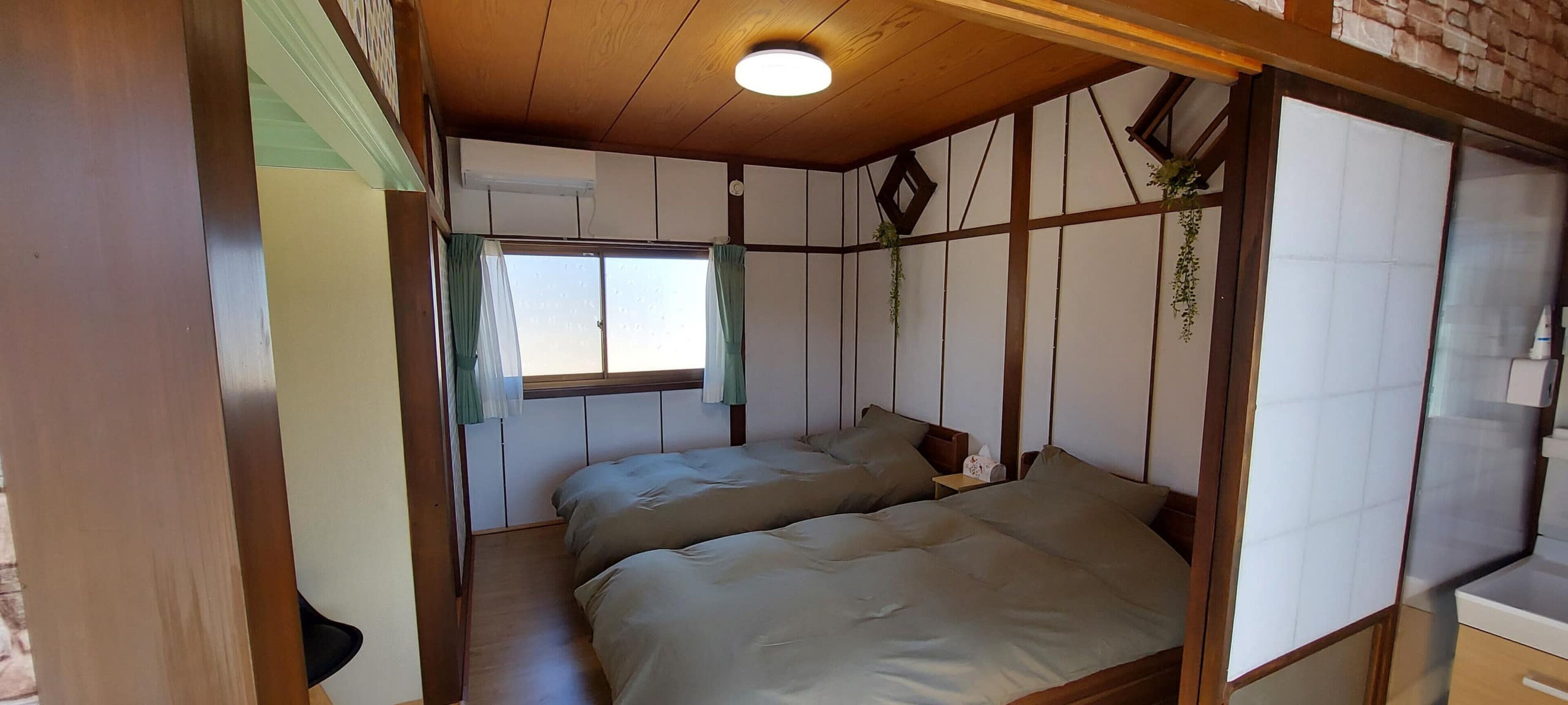 Ein traditioneller japanischer Raum mit Tatami-Böden, zwei Betten, einem Fenster mit Vorhängen und einer Holzkονstruktion an der Decke.