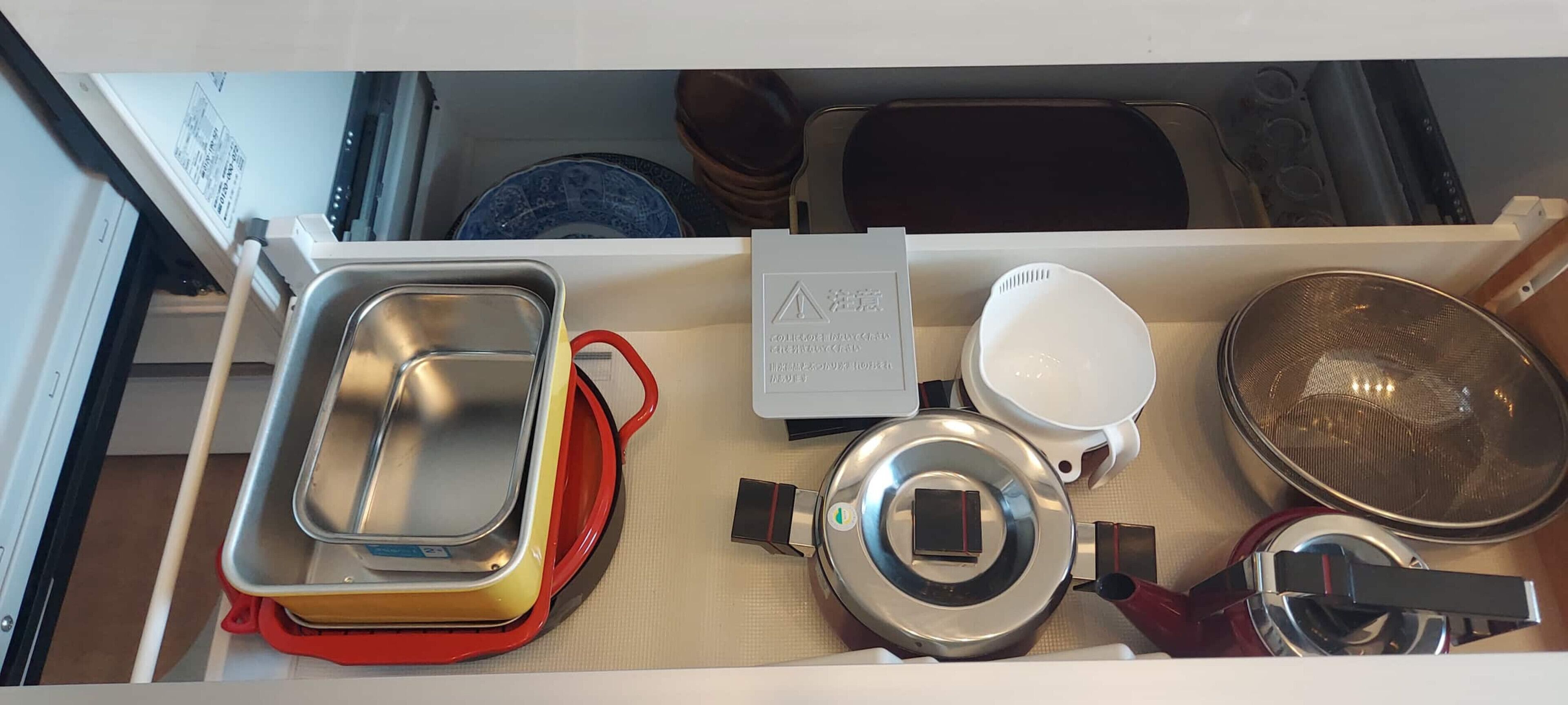 A kitchen drawer with various utensils and containers, including a metal strainer, a white bowl, and a red container.