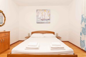 Un lit simple avec du linge de lit et des serviettes blanches, une tête de lit en bois, des lampes de chevet, une commode en bois et un tableau représentant un voilier, accroché au mur.