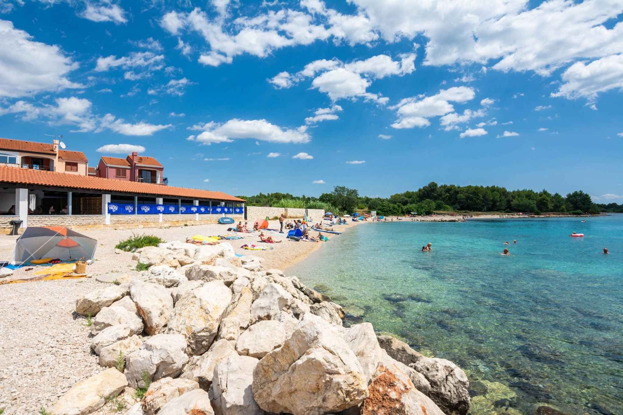 A beach with clear water, a rocky foreground, and a building with a tiled roof.