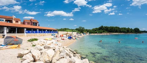 A beach with clear water, a rocky foreground, and a building with a tiled roof.