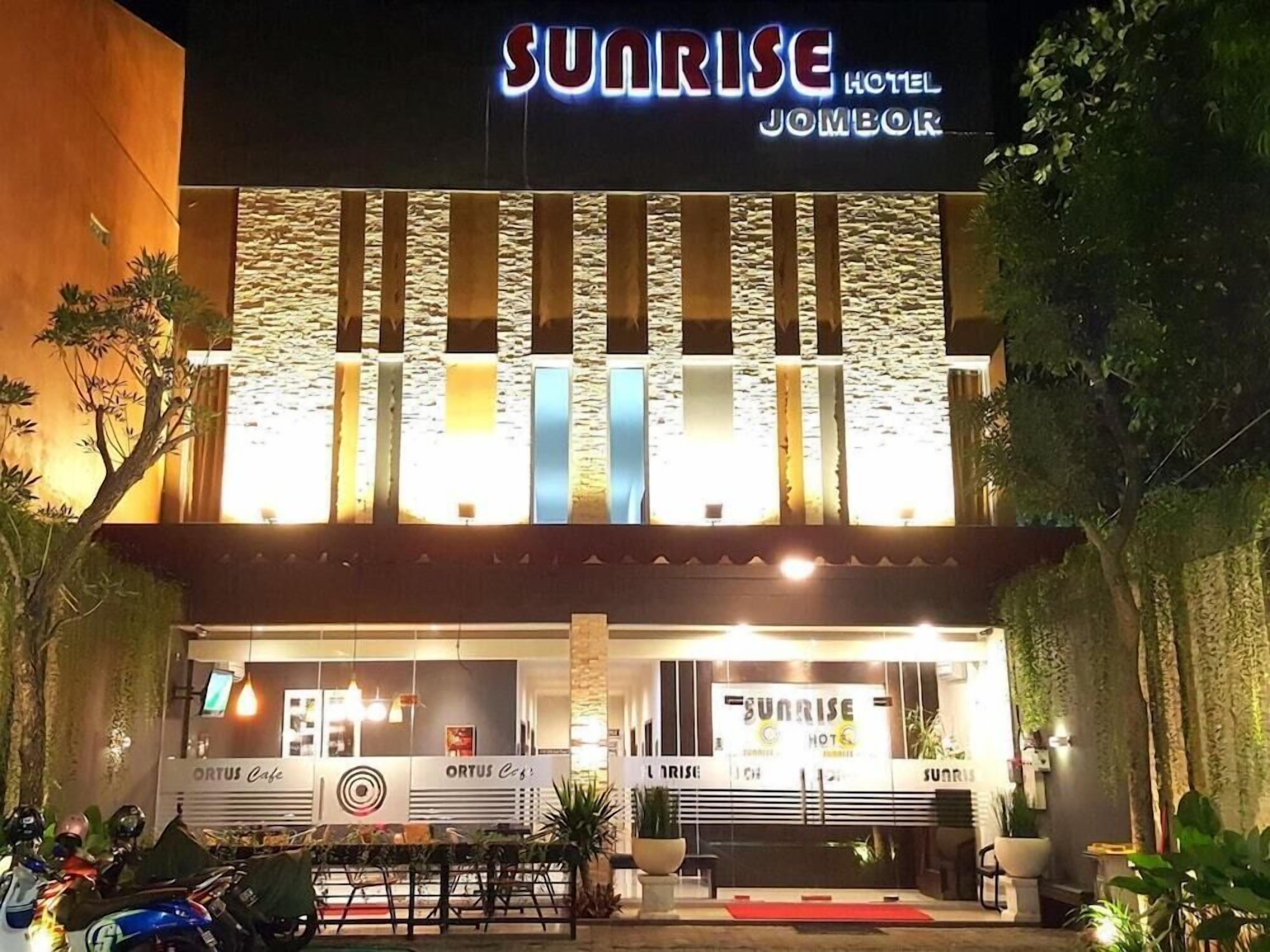 The exterior of Sunrise Hotel Jombor at night with illuminated signage, a visible entrance, and parked motorcycles.