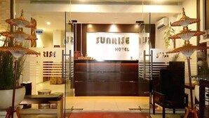 L’entrée d’un hôtel, avec un comptoir d’accueil, une enseigne indiquant « SUNRISE HOTEL » et des lampes décoratives.
