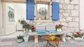 A stone building with a white door, blue shutters, a cushioned bench, potted plants, and a table with fruit. - Holiday home with Wi-Fi and air conditioning (Funtana)