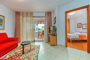 Ein Zimmer mit rotem Sofa, Fernseher, Bett und Blick durch eine Tür.